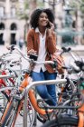 Sorrindo afro-americano fêmea de pé perto de bicicleta estacionada e falando no telefone celular na cidade, enquanto olha para longe — Fotografia de Stock