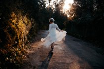 Full body back view of anonymous female wearing white dress walking on rural road among green trees in nature on evening time — Foto stock