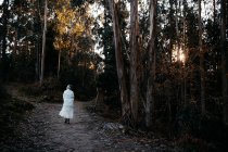 Back view of anonymous female with short hair covered with white blanket strolling on rural path in woods on evening time - foto de stock