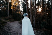 Задний вид анонимной женщины с короткими волосами, покрытыми белым одеялом, прогуливающейся по сельской тропе в лесу в вечернее время — стоковое фото