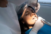 Crop professional dentist in uniform with medical mask drilling tooth of calm woman with help of assistant — Stock Photo