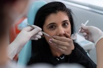 Молода перелякана жінка-пацієнтка дивиться на камеру і прикриває рот руками з лікарем врожаю, який тримає стерильні зубні інструменти — стокове фото