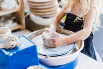 Crop unrecognizable little girl in black apron standing near pottery wheel while shaping clay pot in light workshop — Foto stock