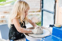 Side view of little girl in black apron standing near pottery wheel while shaping clay pot in light workshop — Stock Photo