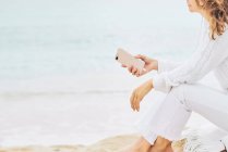 Вид збоку анонімної жінки, що сидить на пляжі біля моря та насолоджується музикою на навушниках та використовує мобільний — стокове фото