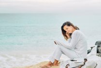 Side view of tranquil female sitting on beach near sea and enjoying music on earphones — Foto stock