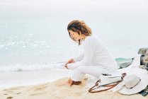Вид сбоку спокойной женщины, сидящей на пляже у моря и наслаждающейся музыкой в наушниках и пользующейся мобильным телефоном — стоковое фото