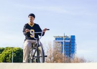 Молодой этнический хипстер мужчина атлет в прохладной одежде сидя на велосипеде bmx, глядя в город в солнечный день — стоковое фото