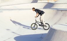 Vista lateral del joven deportista afroamericano realizando trucos en bicicleta de prueba en el parque de skate de la ciudad - foto de stock