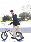 Joven atleta masculino hipster étnico en ropa fresca sentado en la bicicleta BMX mientras mira la cámara en la ciudad en un día soleado - foto de stock