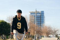 Молодий етнічний хіпстер чоловік-спортсмен в холодному одязі сидить на велосипеді BMX, дивлячись далеко в місто в сонячний день — стокове фото