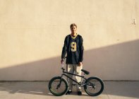 Молодой этнический хипстер мужчина атлет в прохладной одежде сидит на велосипеде BMX, глядя на камеру в городе в солнечный день — стоковое фото