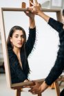 Crop young pensive female in black wear and earring looking at camera against mirror placed on easel — Stock Photo