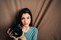 Тысячелетняя женщина с профессиональной фотокамерой сидит на коричневом мятом текстильном фоне и смотрит на камеру дома — стоковое фото