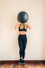 Femme athlétique avec abdos portant haut et leggings debout avec balle noire lourde pour l'entraînement en salle légère pendant l'entraînement — Photo de stock