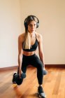 Pieno corpo di donna determinata in abbigliamento sportivo con cuffie ascoltare musica durante l'esercizio affondi con peso a casa — Foto stock