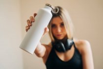 Ruhige Frau mit Kopfhörern in Aktivkleidung, die in die Kamera schaut und Wasser trinkt, während sie nach dem Training zu Hause im Zimmer ruht — Stockfoto