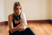 Calm female with headphones wearing activewear looking at camera and drinking water while resting in room after training at home — Stock Photo