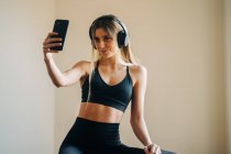 Sportive female in activewear with headphones listening to music and taking self portrait on smartphone while sitting in room after training — Stock Photo