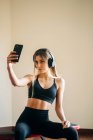 Mulher esportiva em activewear com fones de ouvido ouvindo música e tomando auto-retrato no smartphone enquanto sentado no quarto após o treinamento — Fotografia de Stock