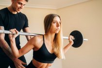 Donna forte in activewear nero facendo esercizio squat bilanciere indietro con l'aiuto di istruttore personale maschile durante l'allenamento a casa — Foto stock