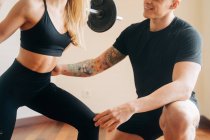Mulher forte em activewear preto fazendo exercício de agachamento de volta barbell com a ajuda de instrutor masculino pessoal durante o treino em casa — Fotografia de Stock