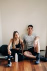 Corpo completo di uomo e donna in abbigliamento sportivo seduti con gambe incrociate sul parquet vicino alla parete con bottiglia d'acqua dopo l'allenamento — Foto stock