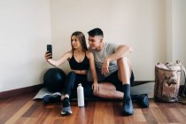 Zufriedene Männer und Frauen in Sportkleidung surfen Handy, während sie auf dem Boden sitzen mit gekreuzten Beinen nahe der Wand mit einer Flasche Wasser — Stockfoto