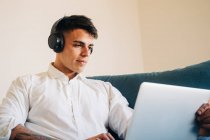 Homem concentrado em fones de ouvido ouvindo música e surfando netbook moderno enquanto sentado no sofá na sala de estar em casa — Fotografia de Stock