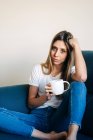 Pensive female with mug of coffee looking at camera and leaning on hand while sitting on couch with bent leg — Stock Photo