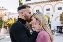 Vista laterale di tenero uomo baciare sorridente donna affascinante in fronte mentre in piedi insieme in città durante la passeggiata — Foto stock