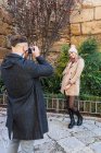 Namorado com câmera de foto tirando foto de namorada encantadora de pé no parque urbano durante o passeio de fim de semana — Fotografia de Stock