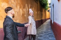 Cheerful couple in love in outerwear holding hands walking along old narrow street while enjoying stroll in city and looking at each other — Stock Photo