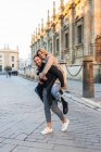 Playful man piggybacking cheerful woman while having fun in city street and looking at camera — Stock Photo