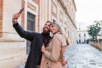 Pareja amante del contenido en ropa de abrigo de moda de pie en la calle y tomando uno mismo tiro en el teléfono inteligente durante el paseo de fin de semana - foto de stock