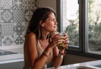 Белая девушка пьет зеленый овощной сок перед своим большим кухонным окном — стоковое фото