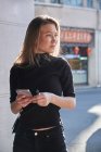 Cultivo de hermosa chica joven rubia usando su teléfono inteligente mirando preocupado con ropa negra - foto de stock