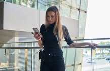 Recadrage de belle jeune fille blonde en utilisant son smartphone regardant inquiet avec des vêtements noirs — Photo de stock