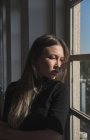 Ritratto di bella ragazza con lunghi capelli biondi, è vicino alla finestra e la luce del sole le illumina il viso. — Foto stock