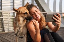 Smiling woman sitting near German Shepherd dog during break in running training and taking self portrait on mobile phone — Stock Photo