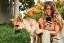 Жінка з довгим волоссям сидить на траві біля німецького улюбленця Шепарда і користується мобільним телефоном у парку. — стокове фото