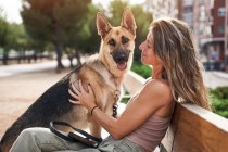 Позитивна жінка - власник обіймає німецького пса Шепарда, сидячи разом на лавці. — стокове фото