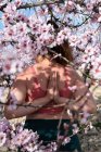 Анонимная женщина в активной одежде, стоящая в позе йоги с руками намасте за спиной, практикуя памятование среди цветущих миндальных деревьев — стоковое фото