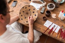 De cima da colheita homem sem rosto sentado à mesa com escovas e esboços de desenho em placa de cerâmica artesanal — Fotografia de Stock