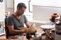 Mann sitzt mit Pinseln am Tisch und zeichnet Skizzen auf handgefertigtem Keramikteller — Stockfoto