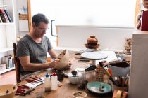 Человек сидит за столом с кисточками и рисует эскизы на ручной керамической пластины — стоковое фото