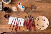 From above of various brushes near painted ceramic plate on wooden table in workshop — Stock Photo