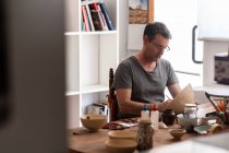Homem sentado à mesa com escovas e desenhos em placa de cerâmica artesanal — Fotografia de Stock
