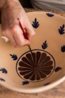 Сверху безурожайный мужчина сидит за столом с кисточками и рисует наброски на керамической тарелке ручной работы — стоковое фото