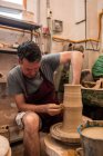Полное тело концентрированного мастера-мужчины в фартуке сидит за столом во время скульптуры с коричневой глиной на бросающем колесе — стоковое фото